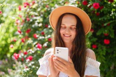 Kahverengi şapkalı ve beyaz elbiseli neşeli genç bir kadın gözleri kapalı gülümsüyor, akıllı telefonuna dalmış kırmızı güller ve yeşil yaprakların arasında..