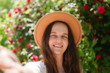 Kahverengi şapkalı neşeli genç bir kadın canlı kırmızı güller ve yeşil yapraklarla güneşli bir selfie çeker..