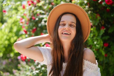 Kahverengi şapkalı neşeli genç bir kadın, kırmızı güllerin ve yeşil yaprakların parlak arka planında saçlarına dokunarak kahkahalarla gülüyor..