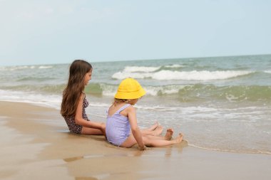 Mayolu iki genç kız, biri uzun saçlı, diğeri sarı şapkalı, kumlu bir sahilde oturup dalgaları seyrediyorlar..