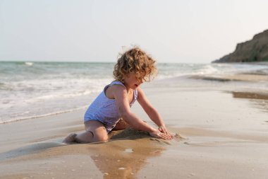 Mayo giymiş kıvırcık saçlı neşeli genç bir kız sahilde kum ve suyla aktif olarak oynuyor..