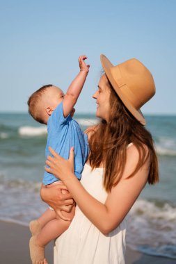 Beyaz elbiseli ve şapkalı gülümseyen bir anne, mutlu bebeğini güneşli bir kumsalda kucağına alıp gökyüzüne uzanır..