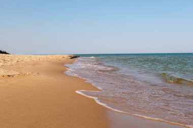 Sahil boyunca uzanan el değmemiş, boş kumlu bir sahil berrak mavi bir gökyüzünün altında kıyıya vuran hafif dalgalarla birlikte..