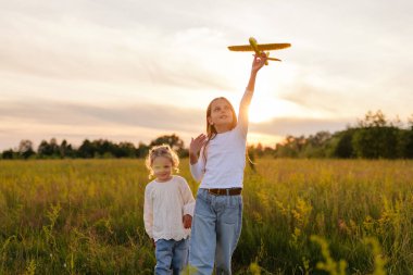 İki genç kız güneşli bir tarlada durur, biri elinde oyuncak bir uçakla, sıcak bir akşam güneşinin tadını çıkarırken..