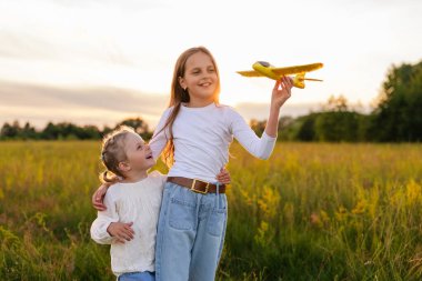 İki neşeli genç kız güneşli bir alanda sarılıyor, biri oyuncak bir uçak tutuyor, çocukluk hayallerini ve sevgisini somutlaştırıyor..