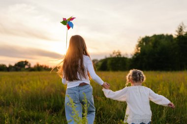 El ele tutuşan iki genç kız, güneş batarken renkli bir fırıldak ile güneşli bir tarlaya doğru yürüyorlar..