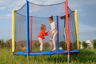 İki oyuncu genç kız, hava bulutlu bir gökyüzünün altında çimenli bir açık alanda güvenli bir ağla canlı bir trambolinin içinde atlayıp el ele tutuşuyorlar..