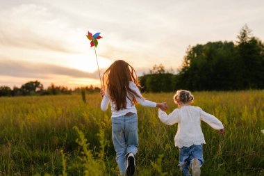 Arkadan görülen iki genç kız, gün batımında güneşten sırılsıklam olmuş bir tarlada el ele koşuyorlar, biri renkli bir fırıldak taşıyor..