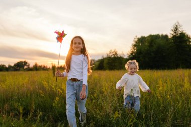 İki neşeli genç kız gün batımında güneşli bir tarlada koşuyor, biri renkli bir fırıldak tutuyor, ikisi de dışarıda altın saatin tadını çıkarıyor..