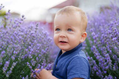 Sarı saçlı ve mavi gözlü neşeli bir bebek, arka planda bir evi olan canlı mor lavanta sıralarının arasında dururken ışıl ışıl gülümsüyor..