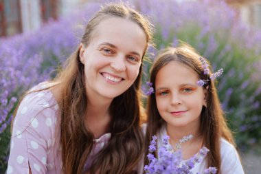 Uzun kahverengi saçlı neşeli bir kadın ve genç bir kız ellerinde lavanta tutarken kameraya gülümserler. Mor çiçekler yumuşak bir arkaplan oluşturur..