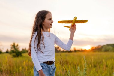 Profilde görünen uzun kahverengi saçlı genç bir kız, yumuşak, güneşli bir gökyüzüne ve ufukta güneş batarken bulanık yeşil bir tarlaya karşı sarı bir oyuncak uçak tutuyor..