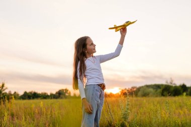 Uzun kahverengi saçlı, beyaz gömlek ve kot pantolon giyen genç bir kız altın bir tarlada elinde sarı oyuncak bir uçakla gün batımına doğru gülümsüyor..