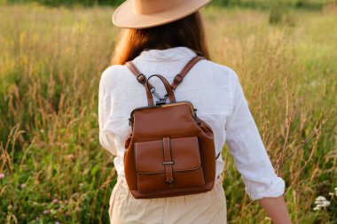 Kahverengi şapkalı, beyaz gömlekli, kahverengi deri çantalı genç bir kadın sırtı kameraya dönük olarak otlak bir alanda duruyor..