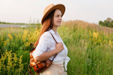 Şapkalı ve sırt çantalı genç bir kadın altın saatinde güneşli, kır çiçekleriyle dolu bir çayıra bakıyor..