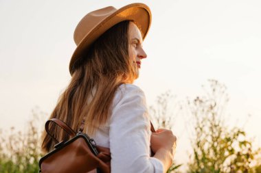 Şapkalı ve beyaz tişörtlü genç bir kadın altın saat boyunca dışarıda duruyor, uzaklara bakarken huzur içinde gülümsüyor, yüzünde güneş ışığı parlıyor..