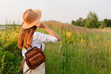 Uzun saçlı, şapka ve sırt çantası takan genç bir kadın, arkasını seyirciye dönük duruyor, güneşli bir çayıra bakıyor..