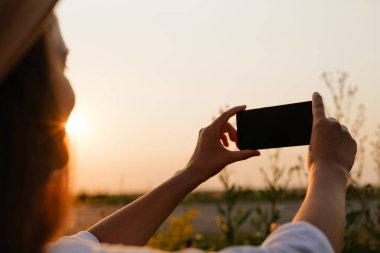 Arkadan bakan bir insan, akıllı telefonuyla bir sahanın üzerinde gün batımını yakalar ve yatay olarak tutar..