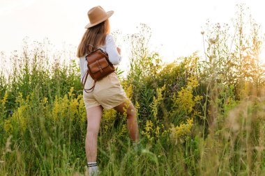 Şortlu, şapkalı ve sırt çantalı genç bir kadın uzun kır çiçekleri ve otların arasında duruyor, altın saat güneşinin tadını çıkarıyor..