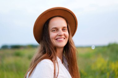 Yeşil bir tarlada gülümseyen kahverengi şapkalı mutlu genç kadın, doğada bir yaz gününün tadını çıkarıyor..