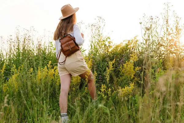 Şortlu, şapkalı ve sırt çantalı genç bir kadın uzun kır çiçekleri ve otların arasında duruyor, altın saat güneşinin tadını çıkarıyor..