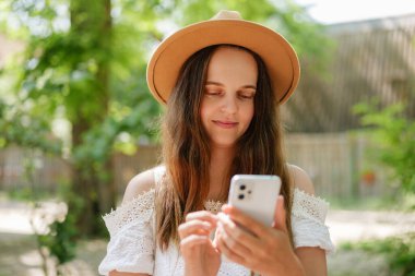 Beyaz omuzlu ve hasır şapkalı genç bir kadın yeşil yaprakların arasında, hafif bir gülümsemeyle akıllı telefonuna bakıyor..