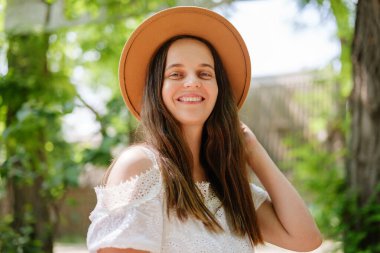 Beyaz omuzlu, saman şapkalı neşeli genç bir kadın kameraya gülümsüyor. Arka planda benekli güneş ışığı ve yeşil yapraklar var..