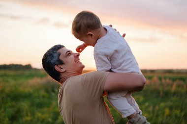 Mutlu bir baba, çimenli bir tarlada gün batımının sıcacık parıltısına gülümseyen küçük çocuğunu havaya kaldırır..