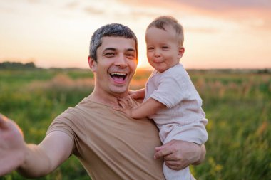 Mutlu bir baba gülümseyen küçük çocuğunu tutar, kameraya bakar, arka planda çimenli bir tarlanın üzerinde sıcak bir günbatımı parıltısıyla..