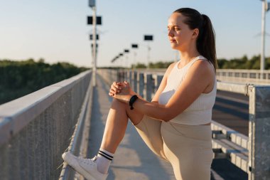 Atletik giysiler içindeki odaklanmış bir kadın, arka planda güneş enerjisiyle çalışan sokak lambaları ve açık gökyüzü olan bir köprüde bacağını esnetiyor..