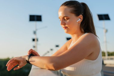 Kablosuz kulaklıklı barışçıl bir kadın, arka planda güneş enerjisiyle çalışan sokak lambalarıyla köprüdeki parmaklıklara dayanan akıllı saatine bakar..
