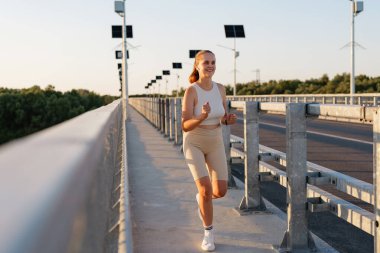 Aktif giyimli neşeli genç bir kadın güneş panelleri yol kenarında ve arka planda yeşil ağaçlarla birlikte gün doğumunda bir köprüde koşuyor..