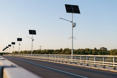 Köprü boyunca güneş enerjisiyle çalışan sokak lambaları ve korkulukların net bir görüntüsü, mesafeye giden bir yol ve parlak bir gökyüzünün altındaki ağaçlar..