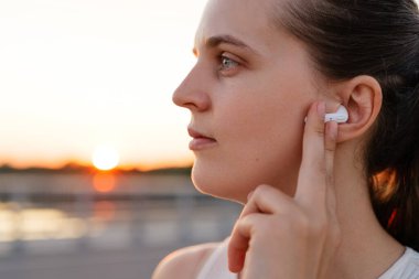 Beyaz kablosuz kulaklığı ayarlayan bir kadının yakın plan profili. Arka planda sıcak bir günbatımı parıltısı var..