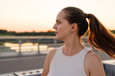 Saçları at kuyruğu olan genç bir kadın, bir köprünün üzerinde gün batımı parlayan ve arka planda bir su kütlesiyle yan tarafa bakıyor..