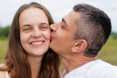 Kısa siyah saçlı bir adam gülümseyen bir kadını yanağından öper. Arka planı bulanıktır. Sevgi dolu bir anı yakalar..