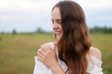 Uzun kahverengi saçlı ve beyaz dantelli gülümseyen genç bir kadın aşağı ve arka planda bulanık yeşil bir tarla ile yan bakıyor.
