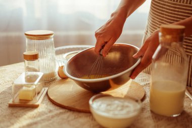 Bir insanın ellerine, tereyağı, kuru gıdalar ve kırsal bir masada sıcak ışık altında görülebilen cam kaplarda bulunan sıvılar gibi diğer pişirme malzemeleri ile birlikte metal bir kasede çırpma malzemeleri gösterilir..