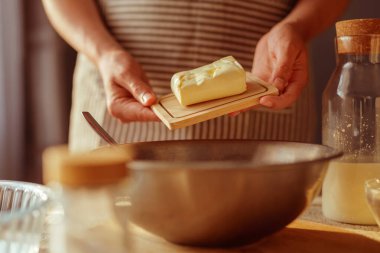 Önlükteki bir insanın elleri, ön planda ve arka planda bulanık olan diğer pişirme malzemeleriyle metal bir karıştırma kabının üzerine yerleştirilmiş küçük tahta bir tahta üzerinde bir kalıp tereyağı tutar..