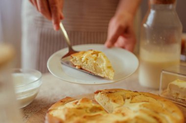 Bir kişi taze pişmiş elmalı turtayı turta sunucusuyla birlikte beyaz bir tabağa koyuyor. Turtanın geri kalanı ve diğer mutfak malzemeleri ön planda ve arka planda bulanıklaşıyor..