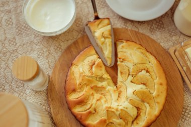 Yeni pişmiş altın elmalı turta, içinden bir dilim dilimlenmiş turta, bir kase kremayla çevrili, ve içinde kuru malzemeler olan cam kavanozlar ve desenli masa örtüsü olan süt..