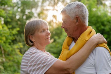 Kapalı, samimi bir açık hava portresi, gri saçlı, birbirine sıcak bir gülümsemeyle bakan, altın güneş ışığıyla yıkanmış, bulanık yeşil yaprakların arasından süzülen, sevgi dolu, yaşlı bir çifti yakalar..