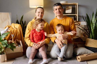 Dört çocuklu gülümseyen bir aile, iki çocuk, bir bebek, yeni bir evde birlikte poz veriyorlar..
