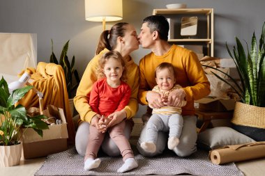 Mutlu ebeveynler öpüşürken, iki küçük çocuk, bir bebek, taşınan kutularla çevrili yeni bir evde kameraya gülümsüyorlar..