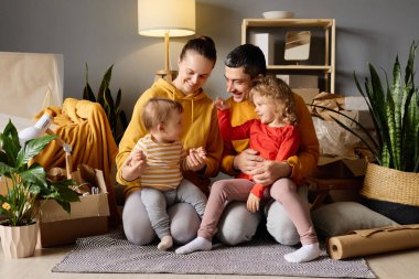 Sevgi dolu dört kişilik aile yeni evlerinde kutularla çevrili bir şekilde yerde otururken, bir bebek ve iki çocuğuyla oynuyorlar..