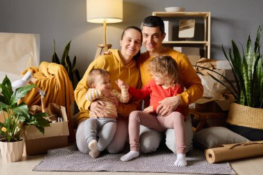 Dört çocuklu mutlu bir aile, bir bebek ve bir bebek, yeni bir evde bir portre için poz veriyorlar, kutuları taşıyorlar, gülümsüyorlar ve kameraya bakıyorlar..