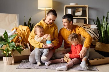 Mutlu ebeveynler iki küçük çocuğuyla oynuyorlar, bir bebek ve bir bebek, yeni evlerinin zemininde oyuncaklarla kutuları taşıyorlar..