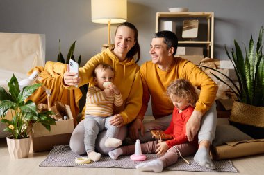 Mutlu ebeveynler yeni evlerinde kutularla çevrili oyuncaklarla oynarken iki küçük çocuklarıyla selfie çekiyorlar..