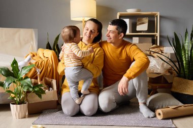 Sevgi dolu ebeveynler yeni bir evde bebekleriyle oynuyorlar, taşınan kutularla çevrili, anne ise bebeği kucağında tutuyor ve gülümsüyor..