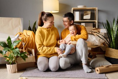 Mutlu genç ebeveynler yeni bir evde gülen bebekleriyle oynuyorlar. Etrafı kutularla çevrili, aileleriyle vakit geçiriyorlar..